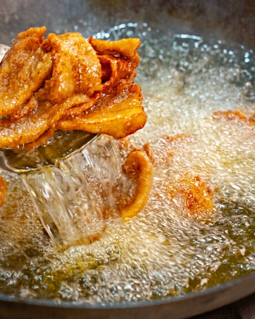 Sacando láminas doradas y curvadas de panceta frita de un wok con aceite hirviendo usando una espumadera.