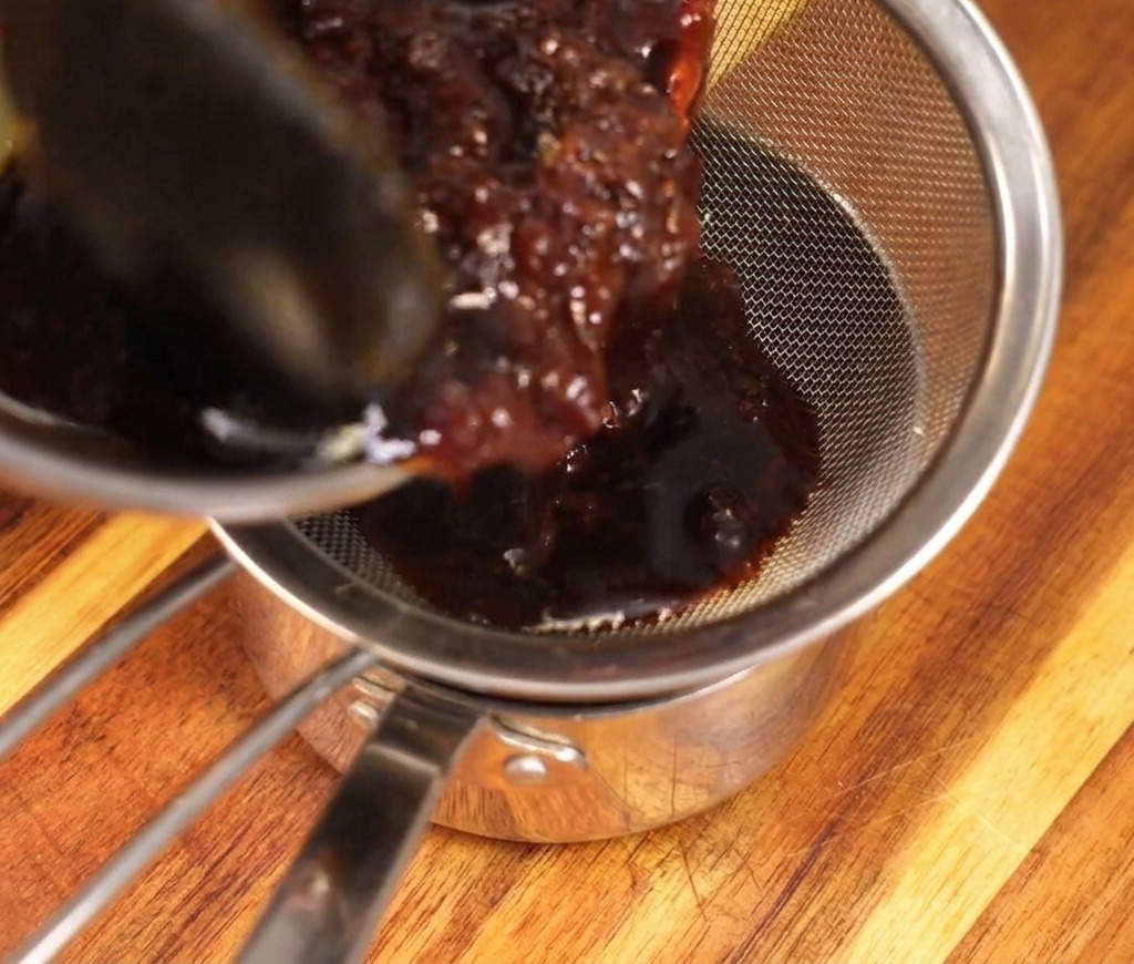 Salsa rica y oscura vertiéndose a través de un colador de malla plateada en un cazo de acero inoxidable, atrapando los copos de bonito empapados.
