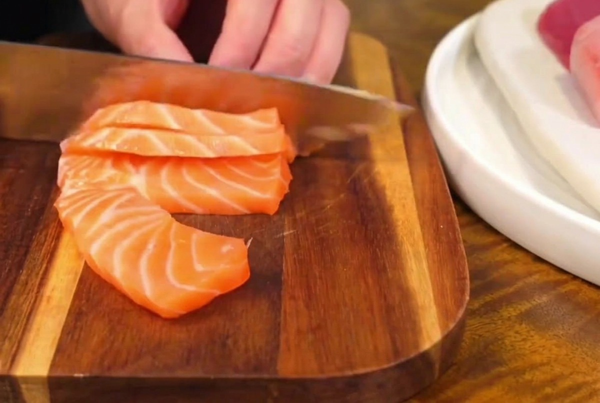 Un cuchillo de cocina afilado cortando un filete de salmón fresco y marmoleado sobre una tabla de madera, creando rebanadas gruesas de sashimi.