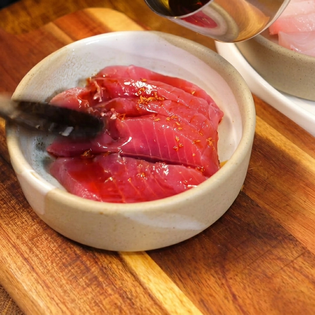 Salsa oscura y sabrosa a base de soja vertiéndose con una cuchara sobre rebanadas de atún crudo de color rojo brillante en un pequeño bol de cerámica rústica.