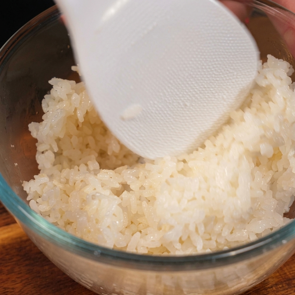 Una paleta de plástico blanco para arroz envolviendo suavemente arroz blanco cocido y sazonado en un bol de vidrio transparente.