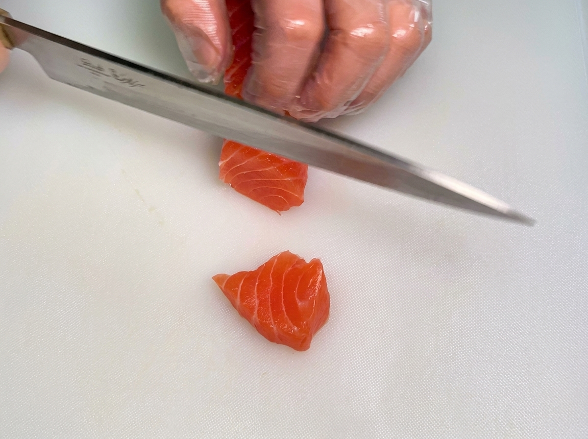 Mano de un chef con guante sosteniendo un bloque de salmón crudo mientras la otra usa un cuchillo para cortarlo en tiras gruesas.