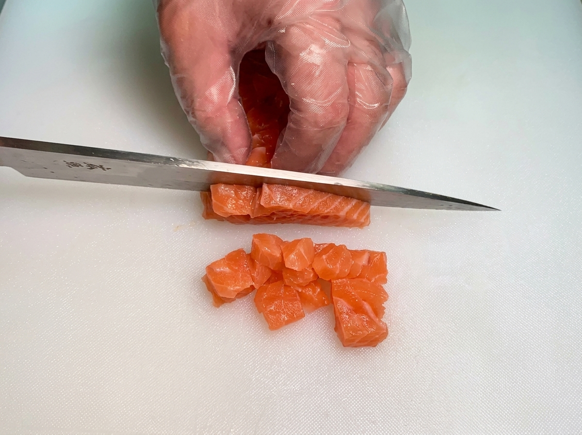 Manos con guantes usando un cuchillo para cortar tiras gruesas de salmón crudo en cubos pequeños uniformes.