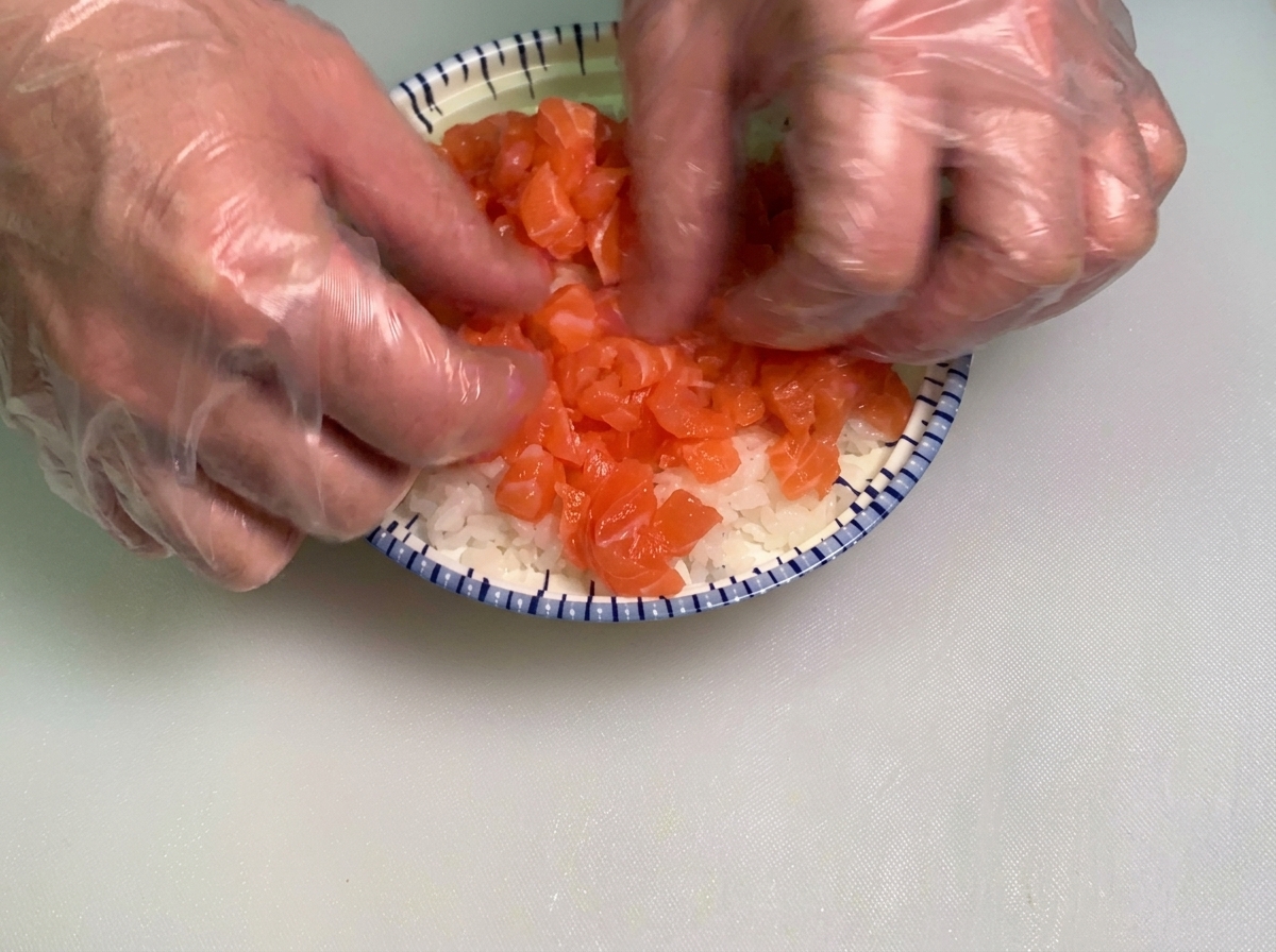Manos con guantes extendiendo suavemente una capa generosa de cubos de salmón crudo sobre una cama de arroz para sushi blanco en un tazón.