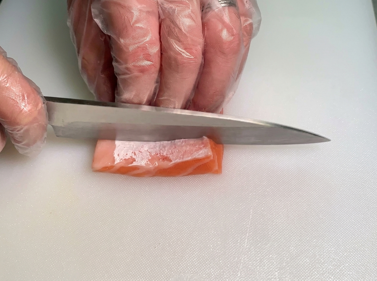 Un chef usando un cuchillo para cortar cuidadosamente un trozo pequeño de ventresca de salmón cruda y grasa en tiras finas.