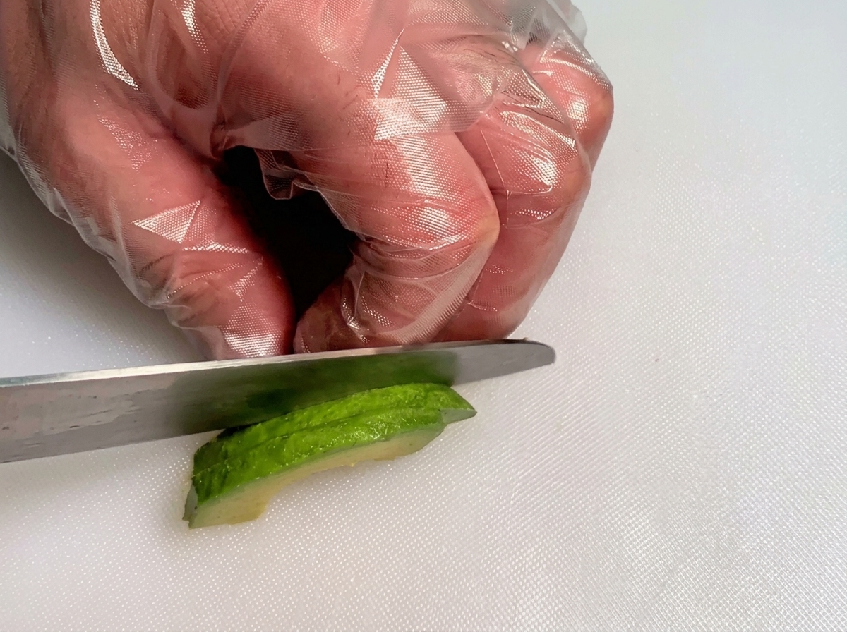 Manos con guantes usando un cuchillo para cortar una rodaja de aguacate verde fresco en cubos pequeños.