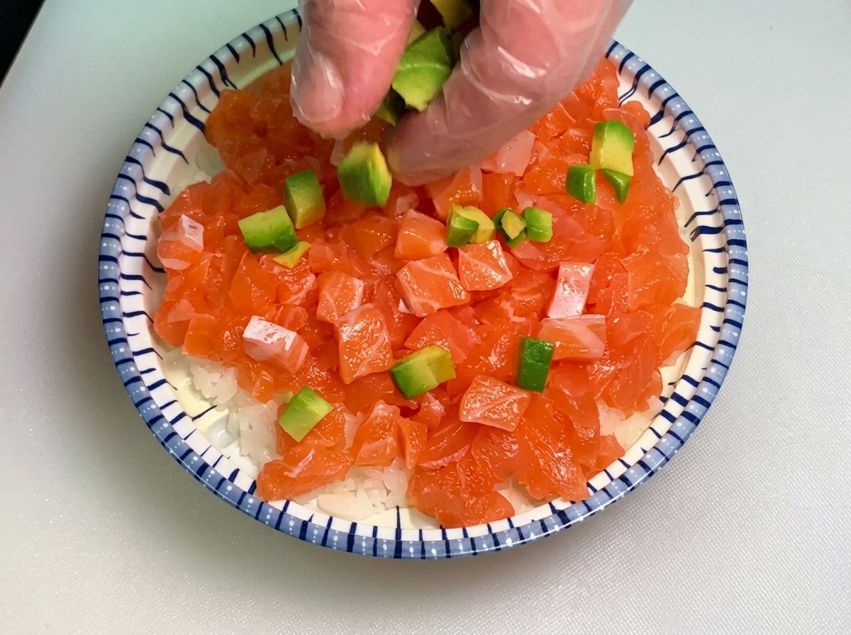 Una mano con un guante transparente esparciendo pequeños cubos de aguacate verde sobre un tazón con capas de salmón en dados y arroz blanco.