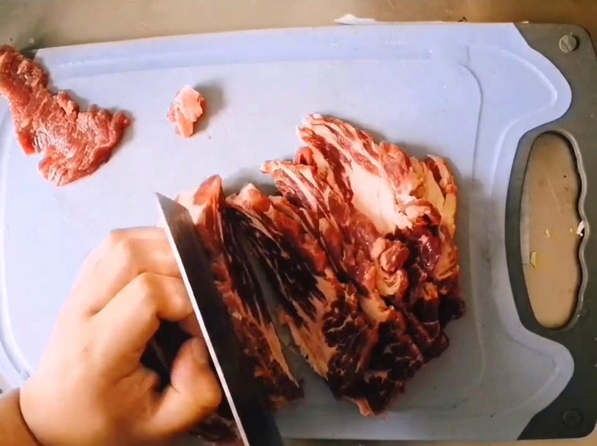 Cortando un trozo de aguja de ternera cruda y veteada en filetes gruesos con un cuchillo afilado sobre una tabla azul claro.