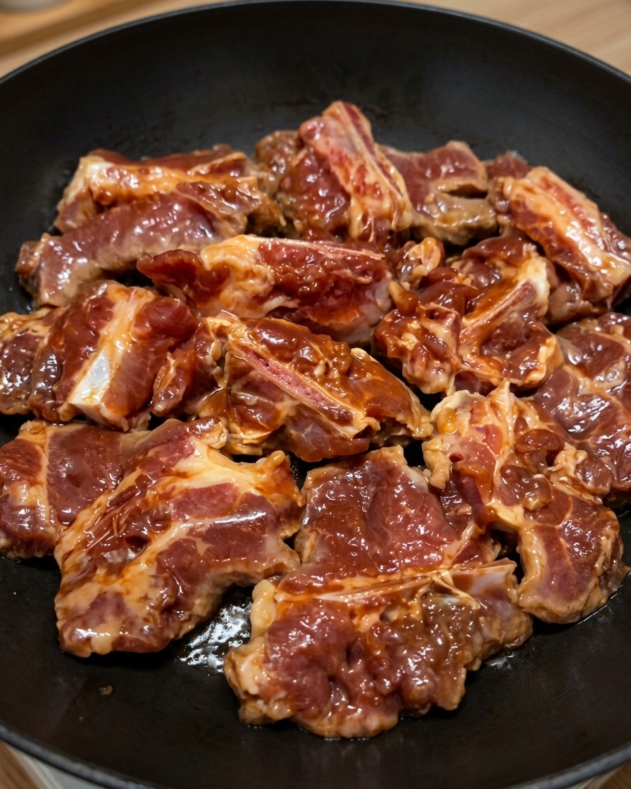 Costillas de cerdo marinadas perfectamente distribuidas en una sola capa sobre una sartén negra plana.