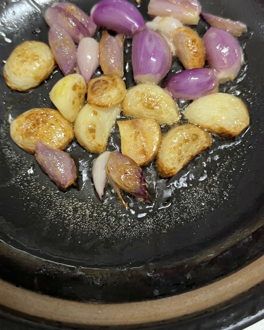 Dientes de ajo enteros y chalotas moradas chisporroteando y dorándose ligeramente en aceite al fondo de una cazuela.