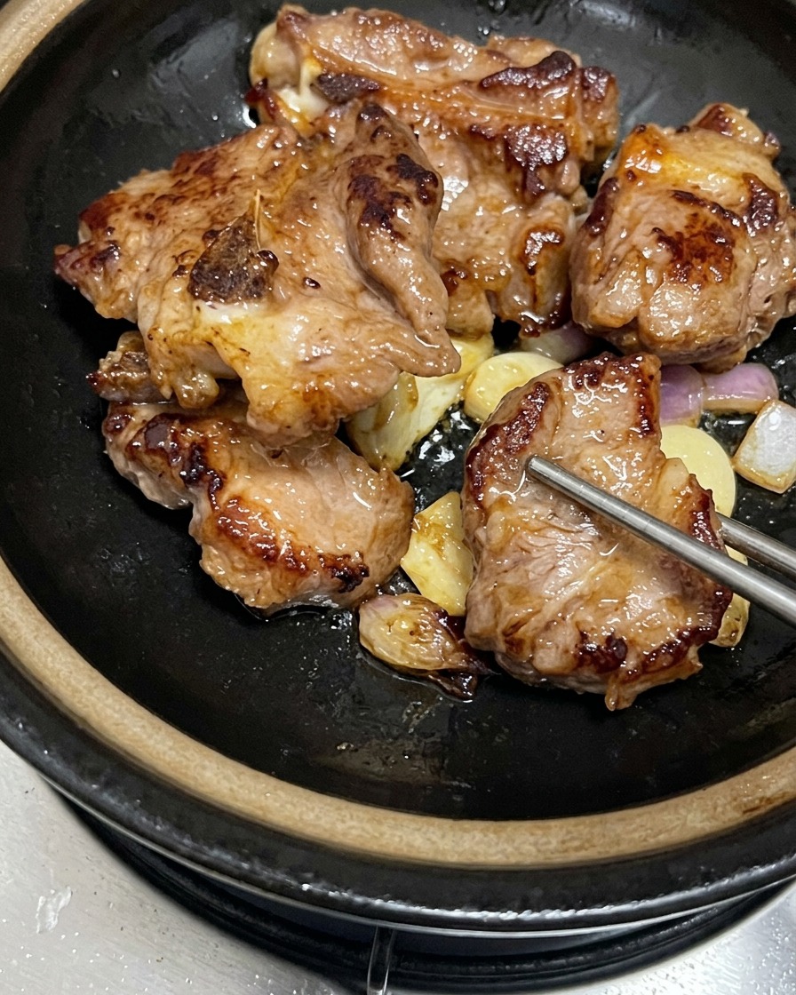 Costillas de cerdo doradas y caramelizadas acomodadas sobre los ajos y chalotas sofritos dentro de una cazuela de barro negra.