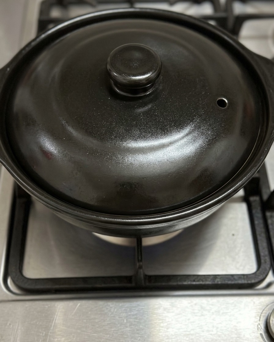 Una tapa de cazuela de barro negra cerrada calentándose sobre la llama azul de una estufa de gas.
