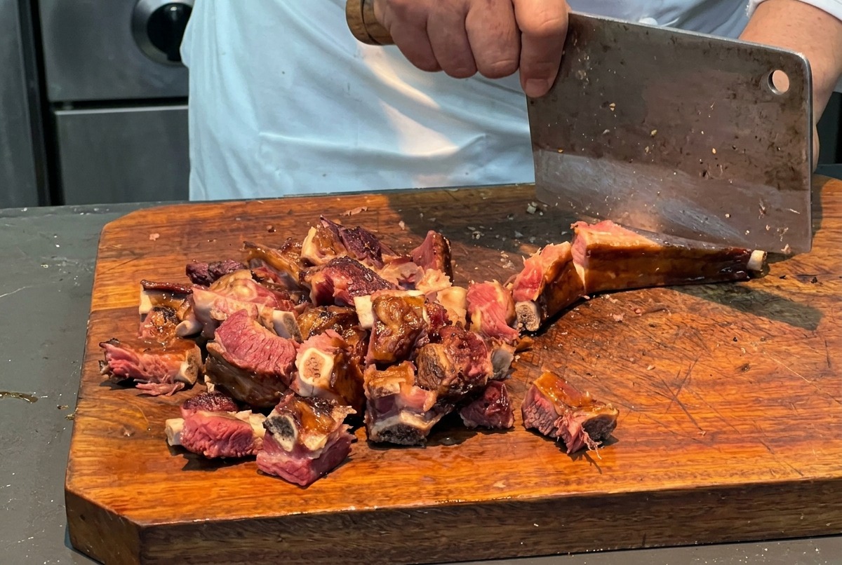 Un chef utilizando un hacha de cocina para cortar un costillar en porciones uniformes de 3 centímetros sobre una tabla de madera.