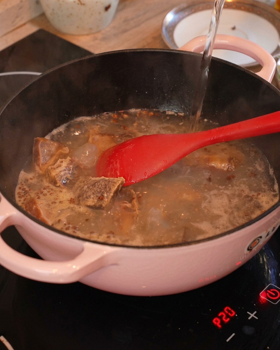 Agua caliente vertiéndose en una olla rosa llena de costillas de cerdo sofritas, jengibre y pimienta de Sichuan.