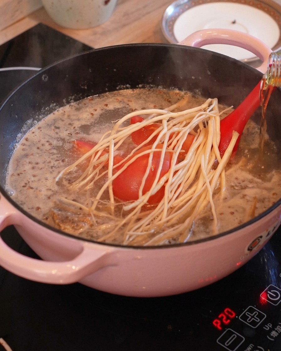 Trozos de tomate y raíces blancas largas en una olla hirviendo con caldo claro, mientras se vierte vino de cocina desde una botella.