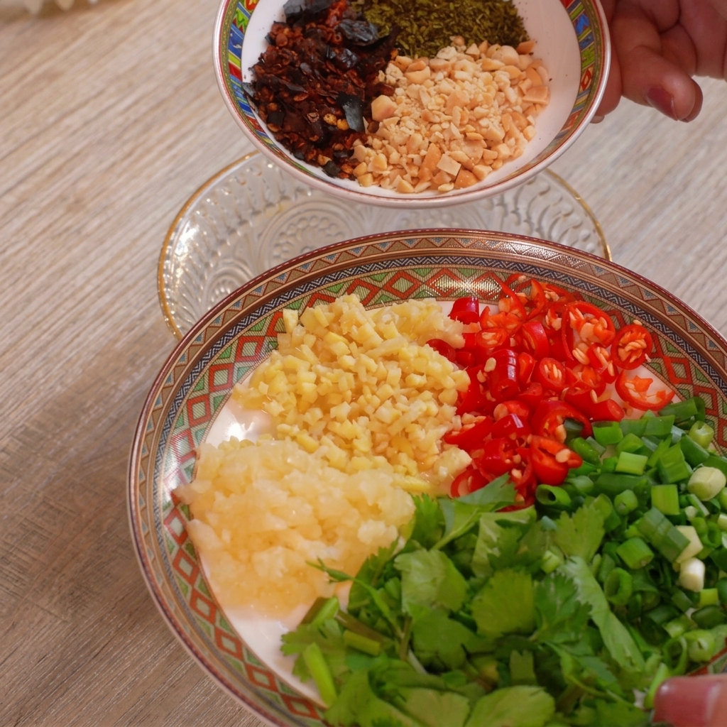 Un plato organizado con montoncitos separados de ajo picado, jengibre picado, chiles rojos troceados, cebolletas y cilantro, con otro bol de cacahuetes y hojuelas de chile encima.