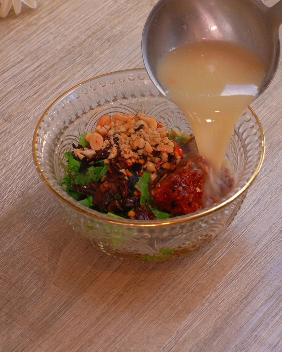 Un cazo de metal vertiendo el rico y lechoso caldo del hot pot en un bol de cristal que tiene capas de hierbas, cacahuetes, chile y tofu fermentado.