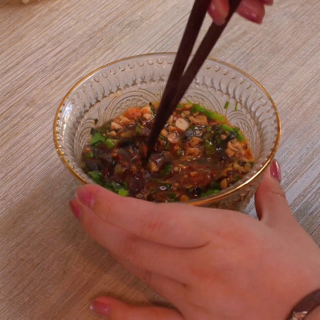 Primer plano de un bol de cristal texturizado lleno de coloridos ingredientes para la salsa como cebolletas verdes, cacahuetes triturados y hojuelas de chile rojo siendo mezclados a fondo con palillos oscuros.