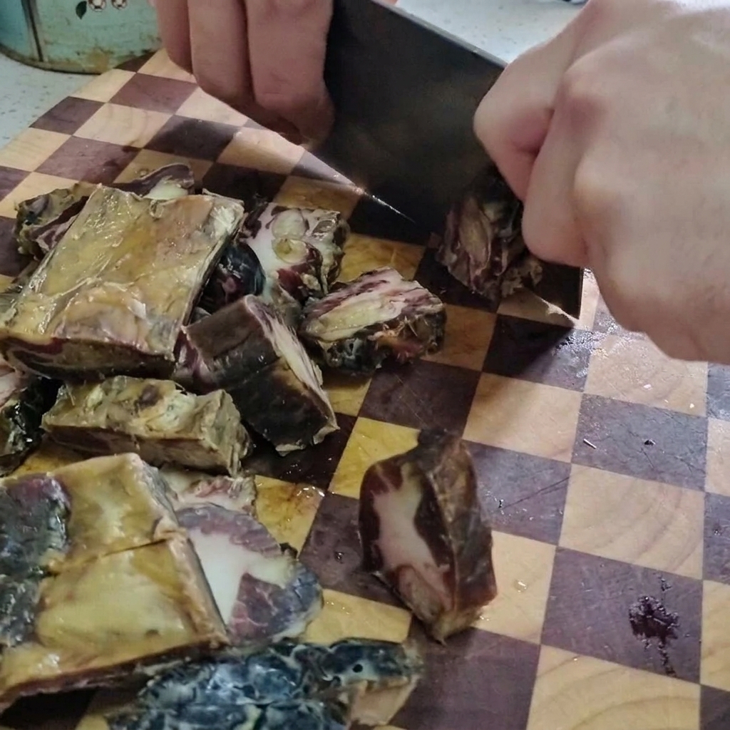 Un plano detalle de una persona usando un hacha de cocina grande para cortar costillas de cerdo curadas en trozos pequeños sobre una tabla de cortar de madera.