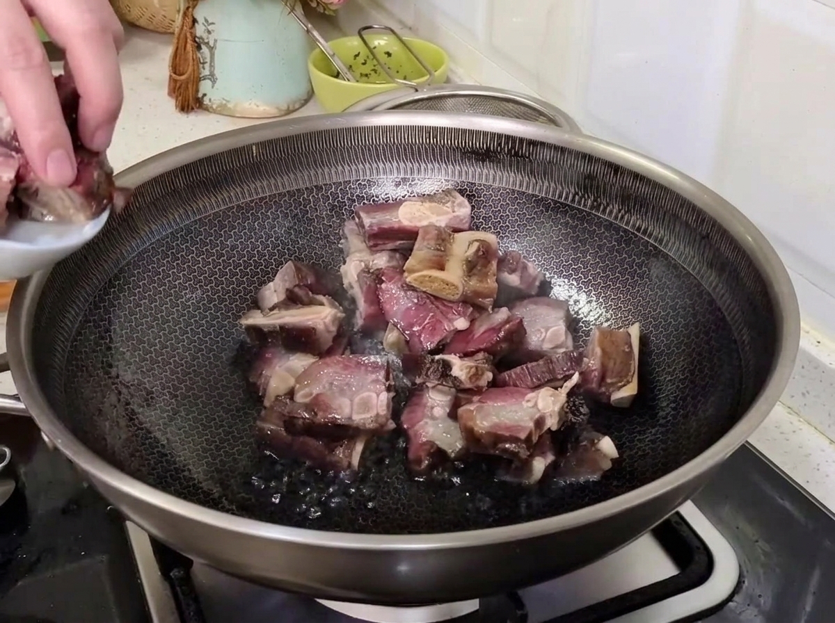 Una mano pasando costillas de cerdo curadas escurridas desde un plato pequeño a un wok caliente para saltearlas.