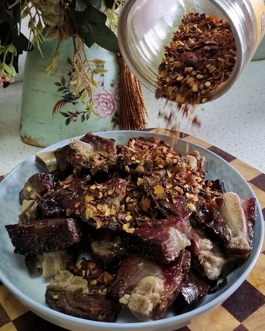 Copos de chile rojo cayendo desde un frasco de vidrio sobre una montaña de costillas de cerdo curadas cocinadas en un plato.