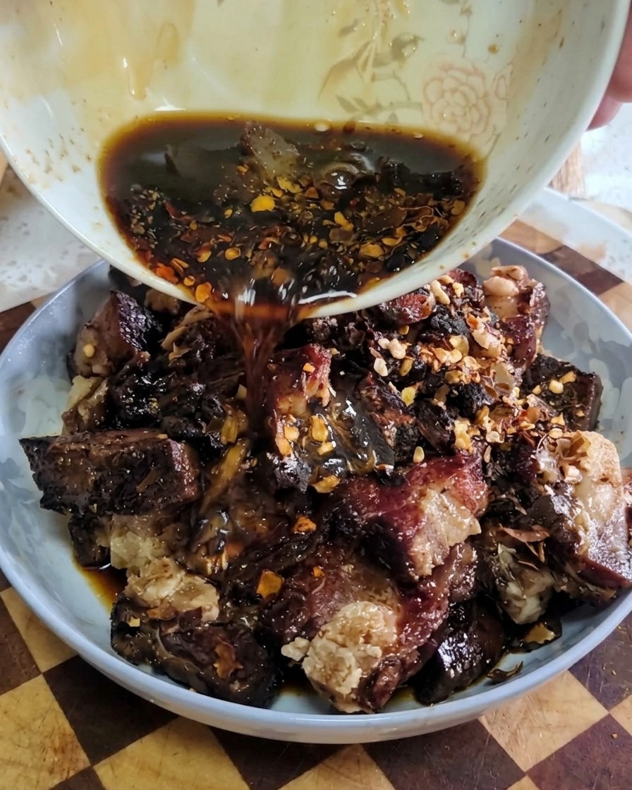 Un líquido oscuro y sabroso cayendo desde un pequeño cuenco blanco sobre un plato de costillas de cerdo curadas y sazonadas.