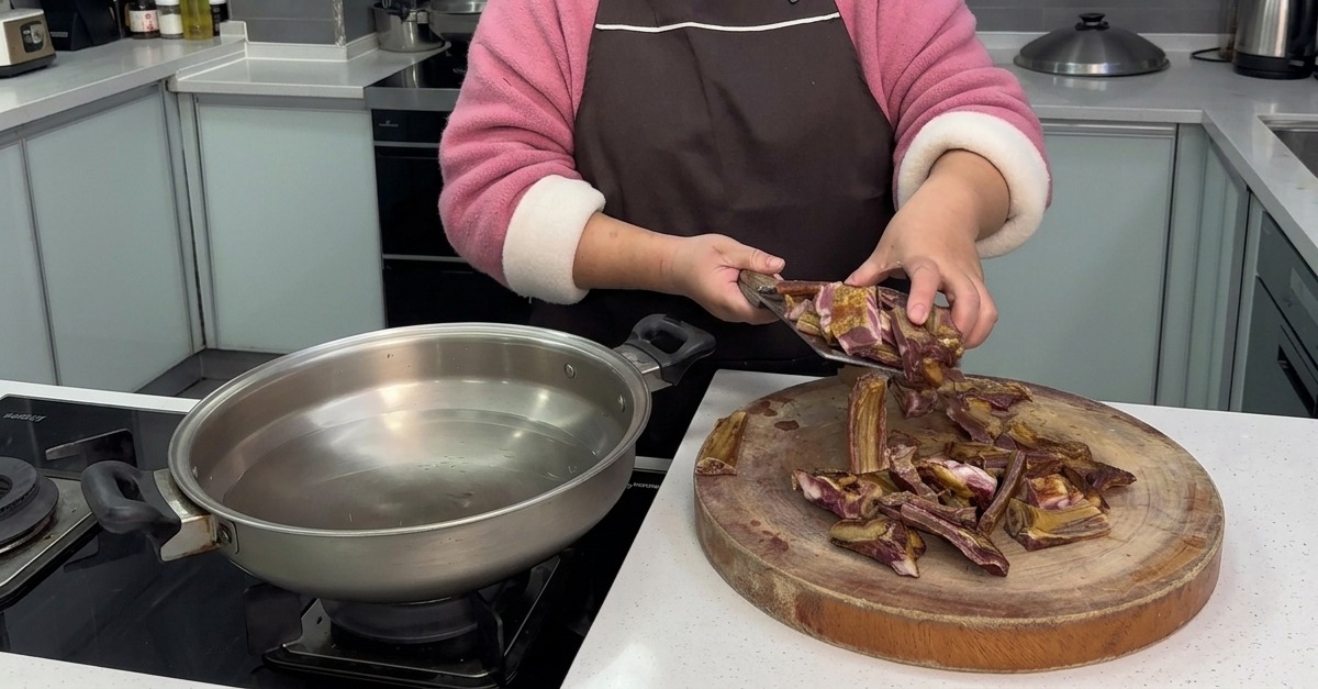 Una persona pasando trozos recién cortados de costillas de cerdo curadas desde una tabla de madera a una cazuela de acero inoxidable con agua fría.