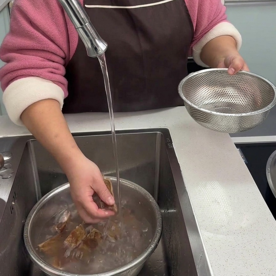 Manos lavando a fondo trozos de costillas de cerdo curadas blanqueadas bajo el chorro de agua fría en el fregadero de la cocina.