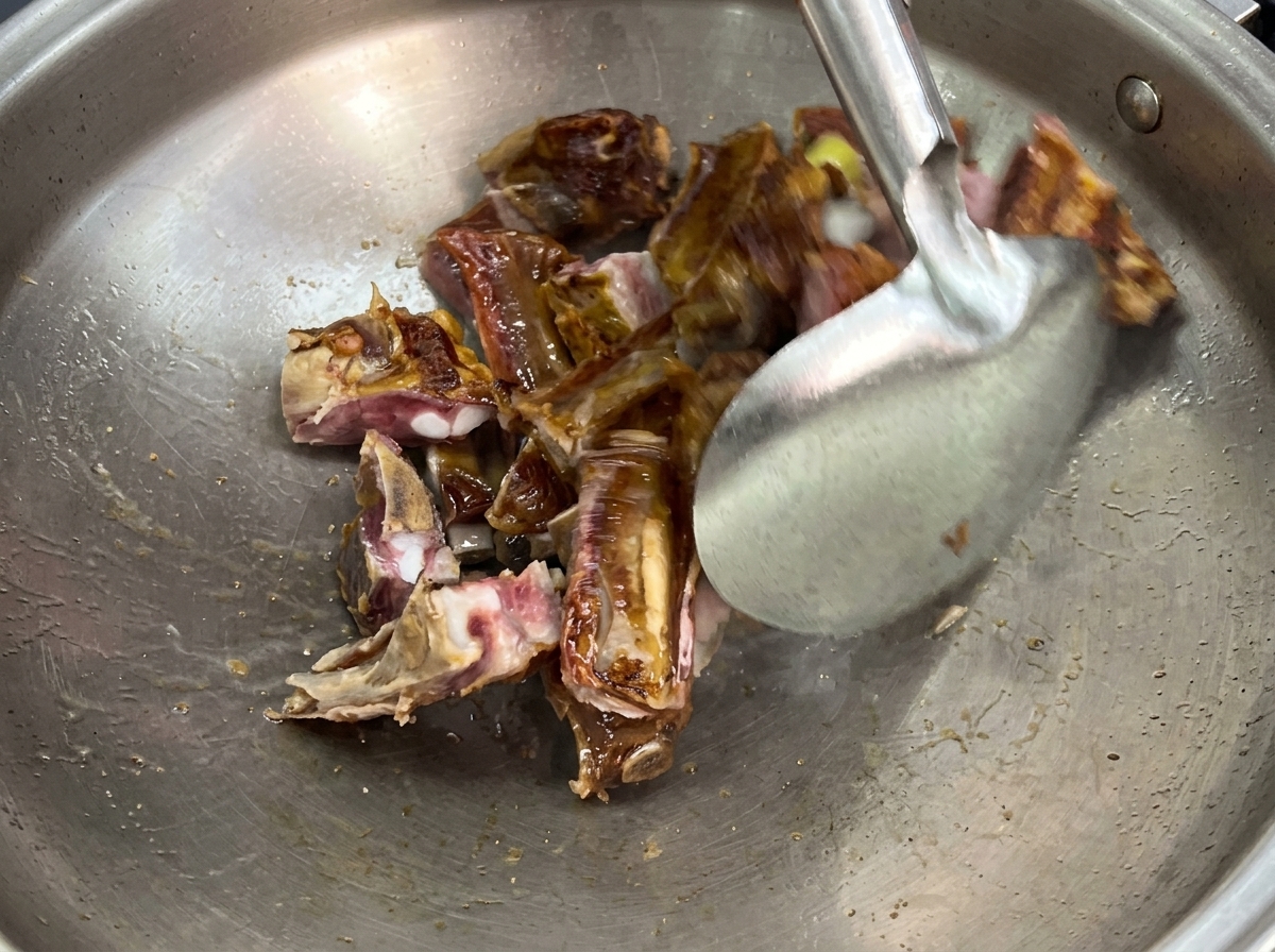 Una espátula de metal salteando trozos de costillas de cerdo curadas y rodajas de jengibre dentro de un wok grande de acero inoxidable.