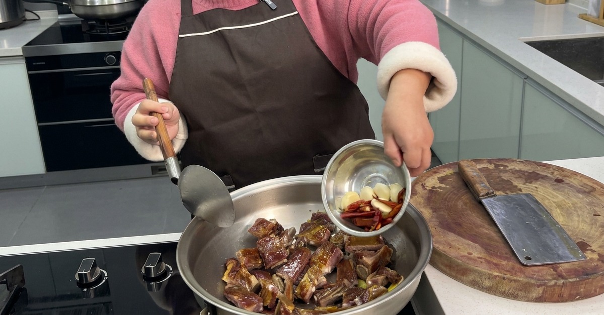 Una persona vertiendo dientes de ajo enteros y chiles rojos secos desde un pequeño cuenco de metal a un wok con costillas curadas salteadas.