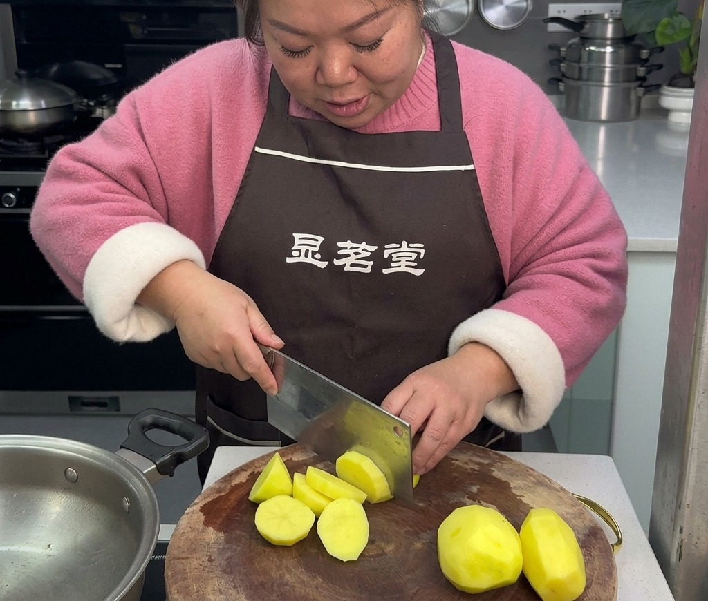 Una mujer con delantal marrón utiliza un cuchillo de cocina grande para cortar patatas amarillas peladas en trozos irregulares sobre una tabla de cortar de madera.