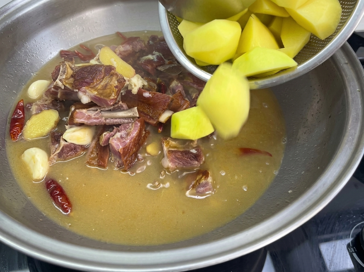 Vertiendo trozos de patata dorados desde un colador a una sartén grande de acero inoxidable con caldo hirviendo, costillas de cerdo curadas, ajo y chiles rojos.