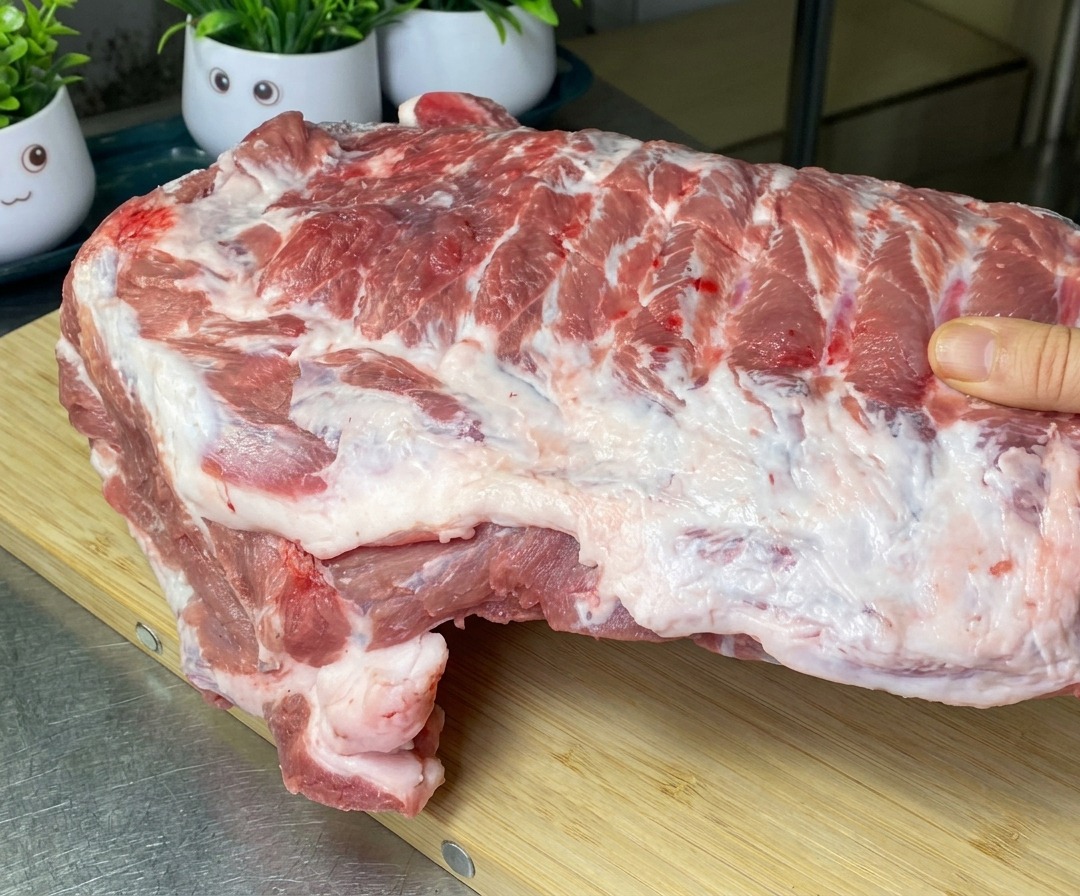 Una gran tira de costillas de cerdo crudas sobre una tabla de cortar de madera, mostrando los cortes iniciales entre los huesos.