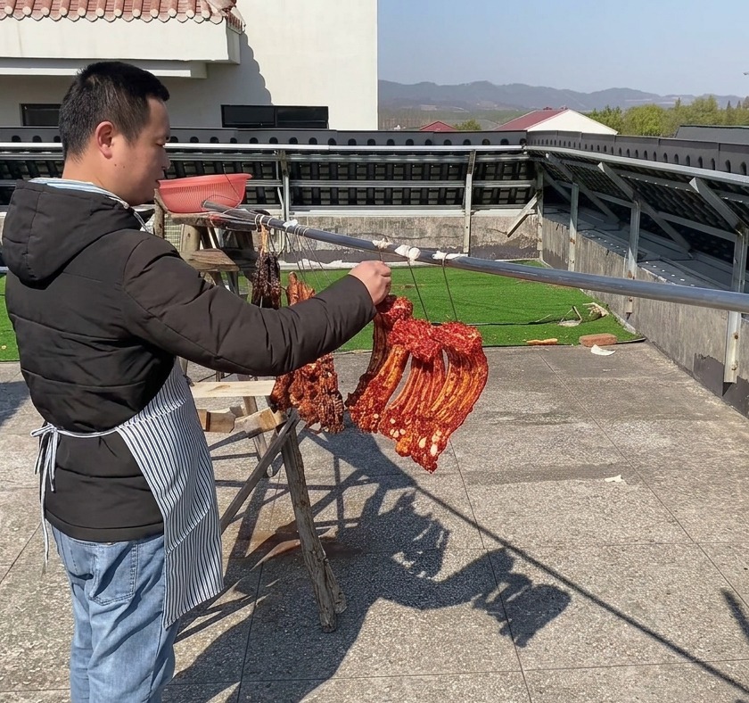 Tiras de costillas de cerdo de color rojo intenso fuertemente condimentadas colgando unas junto a otras en una larga barra metálica exterior para secarse al aire bajo un cielo despejado.