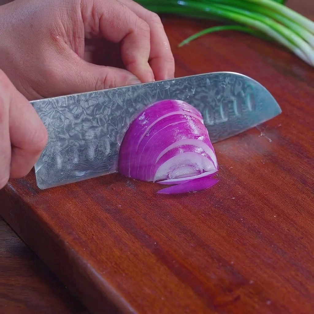 Un cuchillo de chef cortando una cebolla morada en tiras finas sobre una tabla de cortar de madera con cebollas verdes visibles al fondo.