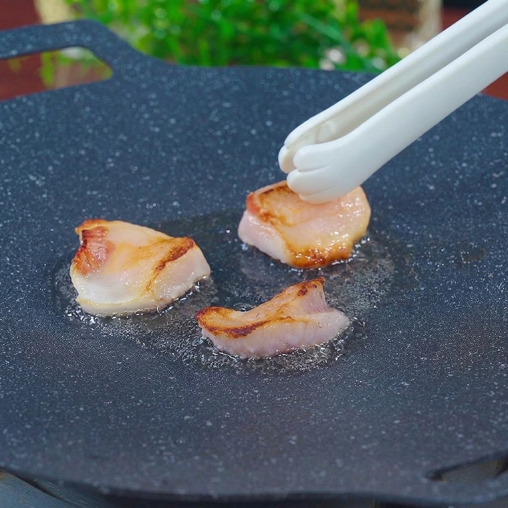 Unas pinzas blancas con forma de pata frotando trozos de grasa de cerdo en una plancha negra caliente para derretir la manteca.