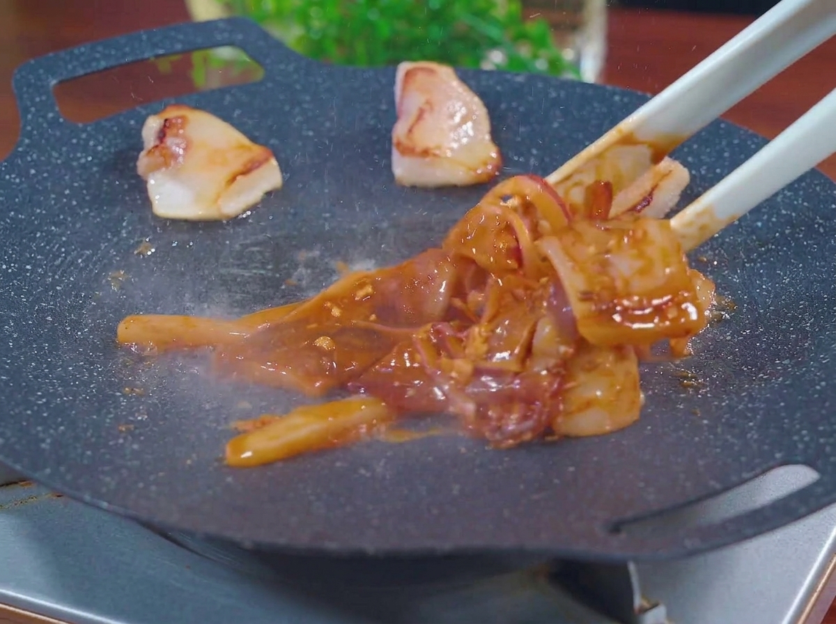Unas pinzas colocando trozos de calamar rojo marinado y rodajas de cebolla en una plancha negra caliente y humeante junto a trozos de grasa de cerdo.