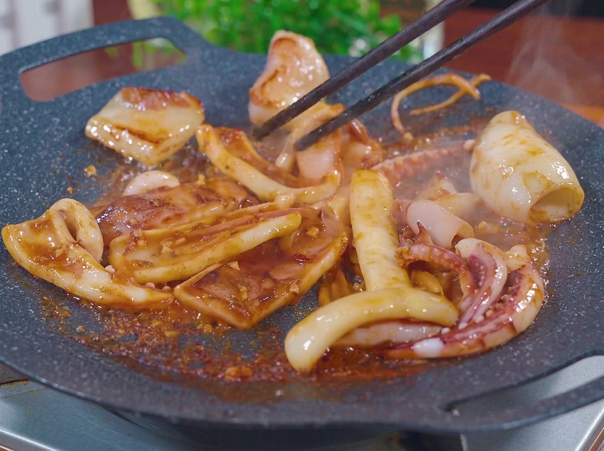 Trozos de calamar salteados con palillos en una plancha caliente, burbujeando en un marinado de barbacoa espeso de color marrón rojizo con vapor subiendo.