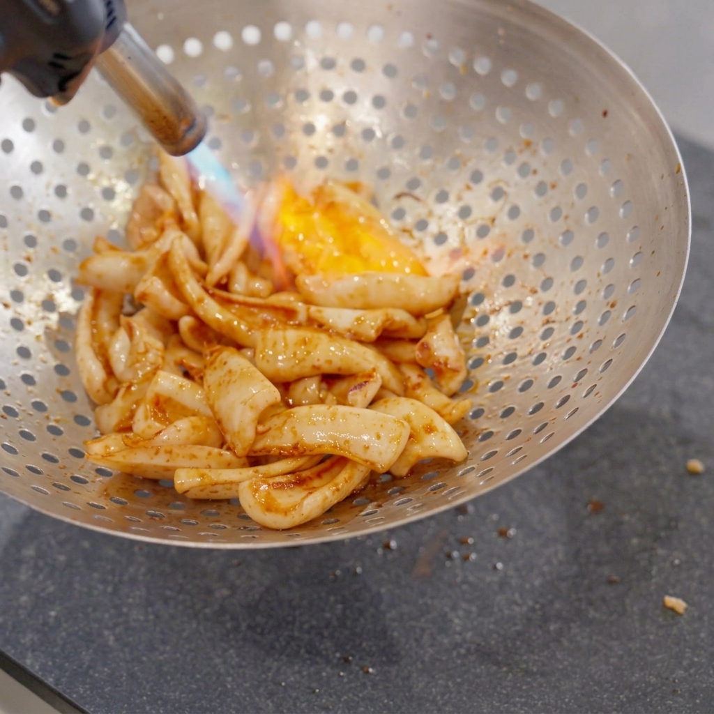 Trozos de calamar frito dentro de un colador metálico perforado grande siendo chamuscados por una llama azul y naranja de un soplete de cocina de mano.