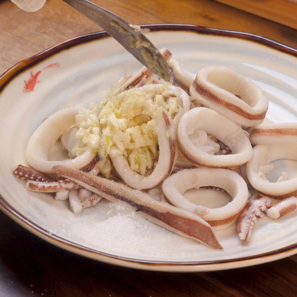 Anillos de calamar blanco escaldados dispuestos en un plato, cubiertos con una montaña de ajo picado y una pizca de sal y azúcar a un lado.
