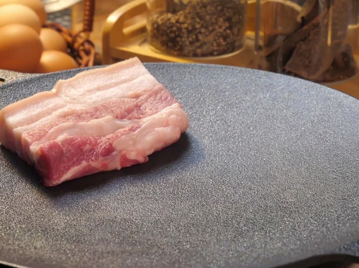Lonchas gruesas de panceta de cerdo cruda colocadas sobre una plancha caliente para dorarse.