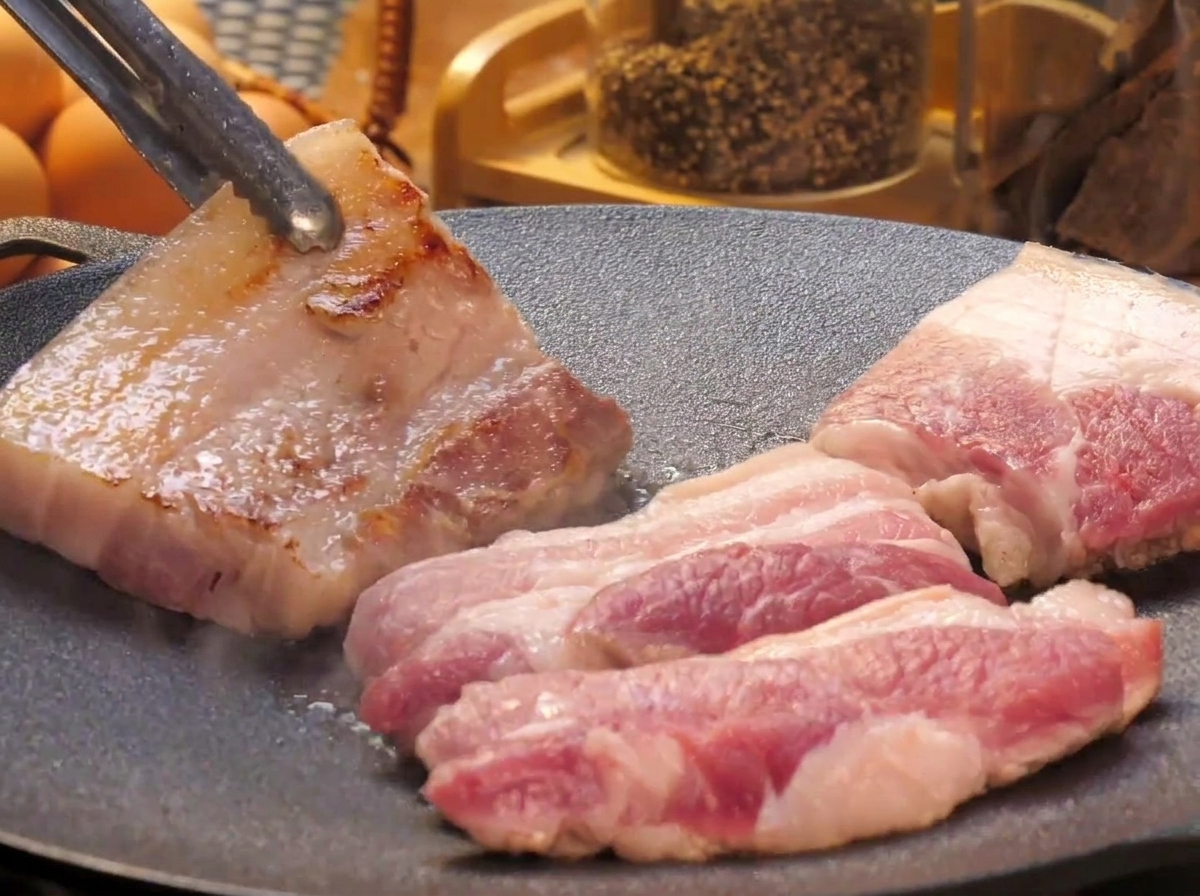 Lonchas gruesas de panceta cocinándose en una plancha, con un trozo al que se le da la vuelta con unas pinzas de metal para mostrar su superficie dorada.