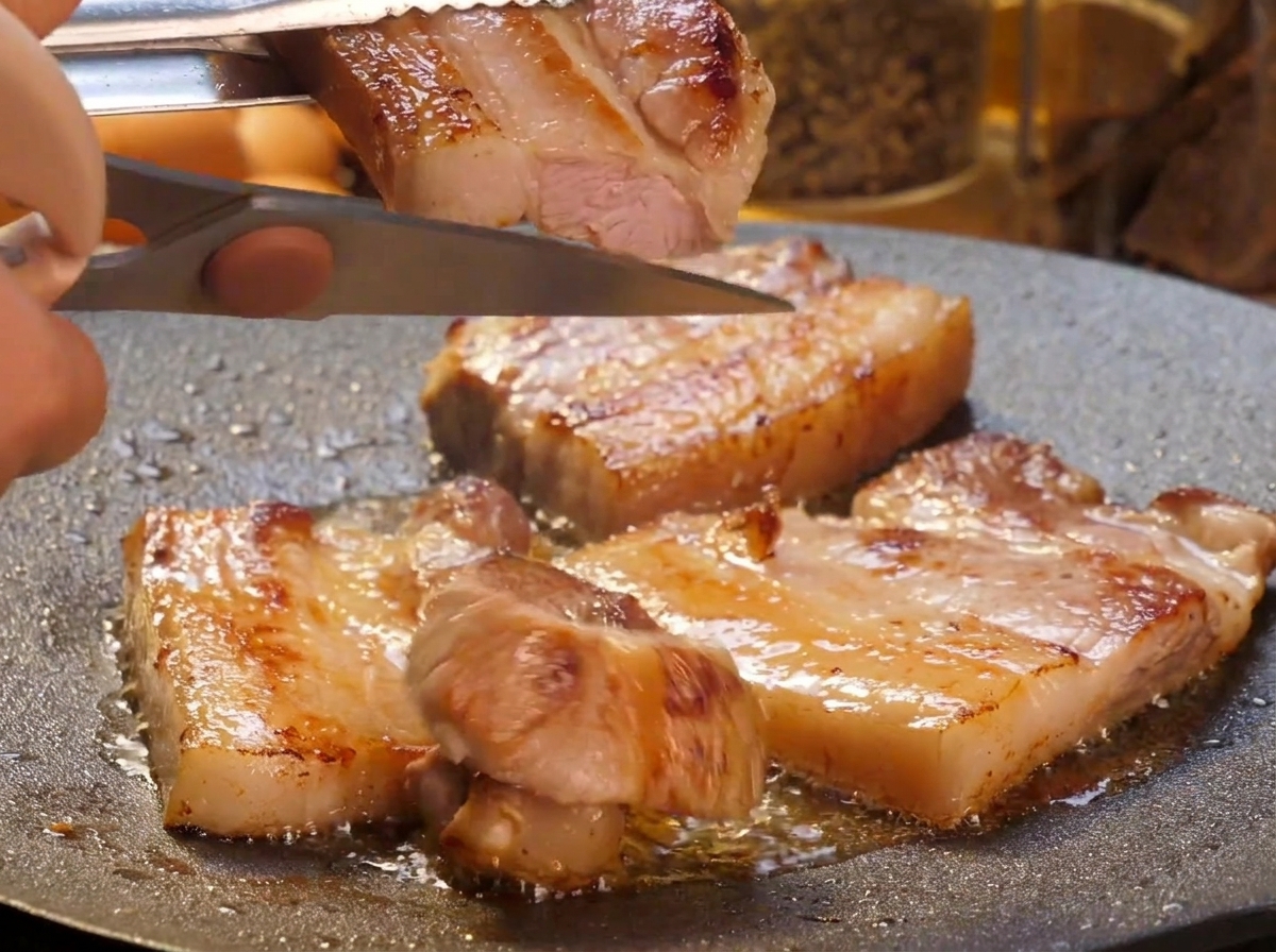 Una mano usando tijeras de cocina para cortar lonchas de panceta doradas en trozos más pequeños sobre una plancha caliente.