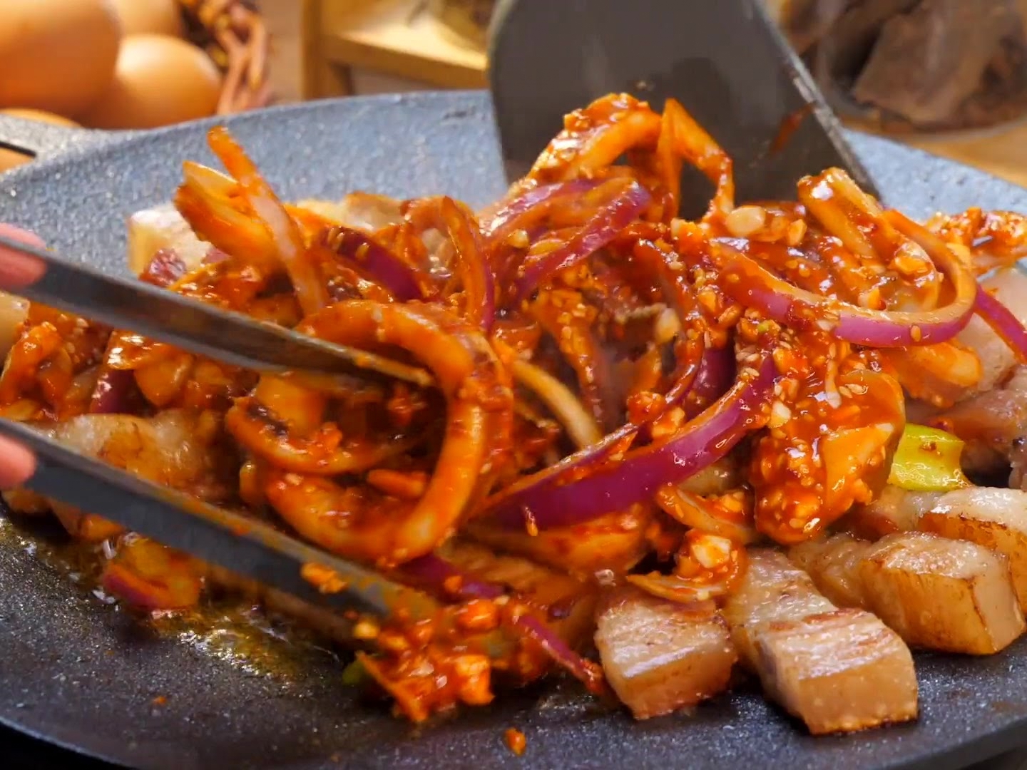 Una espátula y unas pinzas salteando anillos de calamar marinados y trozos de panceta con cebolla morada en una plancha caliente.