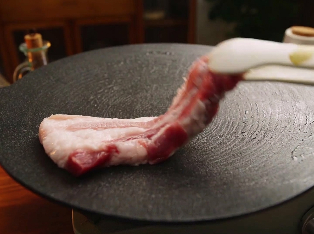 Una tira gruesa de panceta de cerdo cruda colocándose con unas pinzas sobre una plancha de hierro negra y caliente.