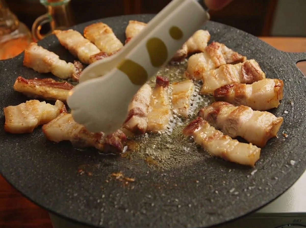 Unas pinzas acomodando trozos de panceta de cerdo a la parrilla en un patrón circular alrededor del borde de una plancha de hierro fundido.