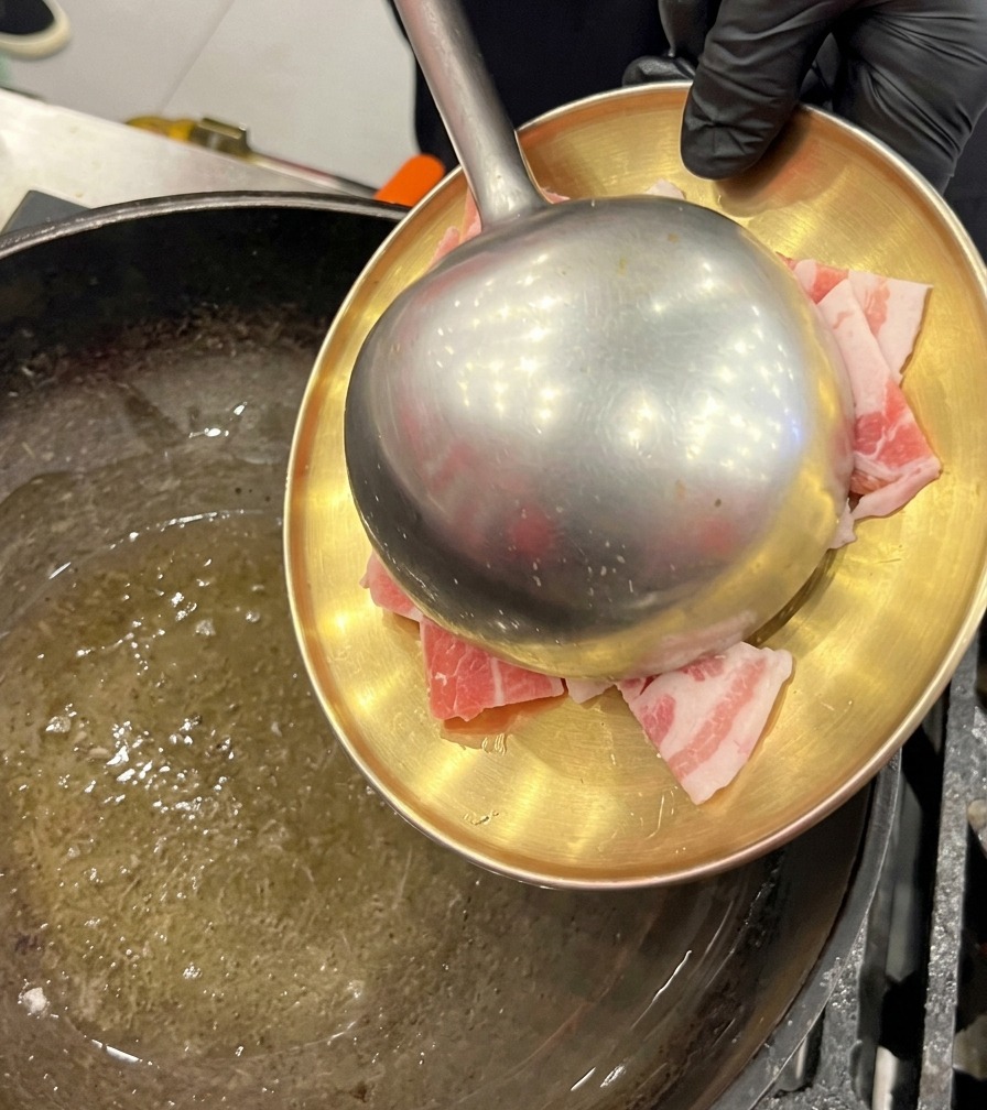 Tiras de panceta cruda siendo pasadas desde un plato dorado hacia un wok con aceite usando un cucharón metálico grande.