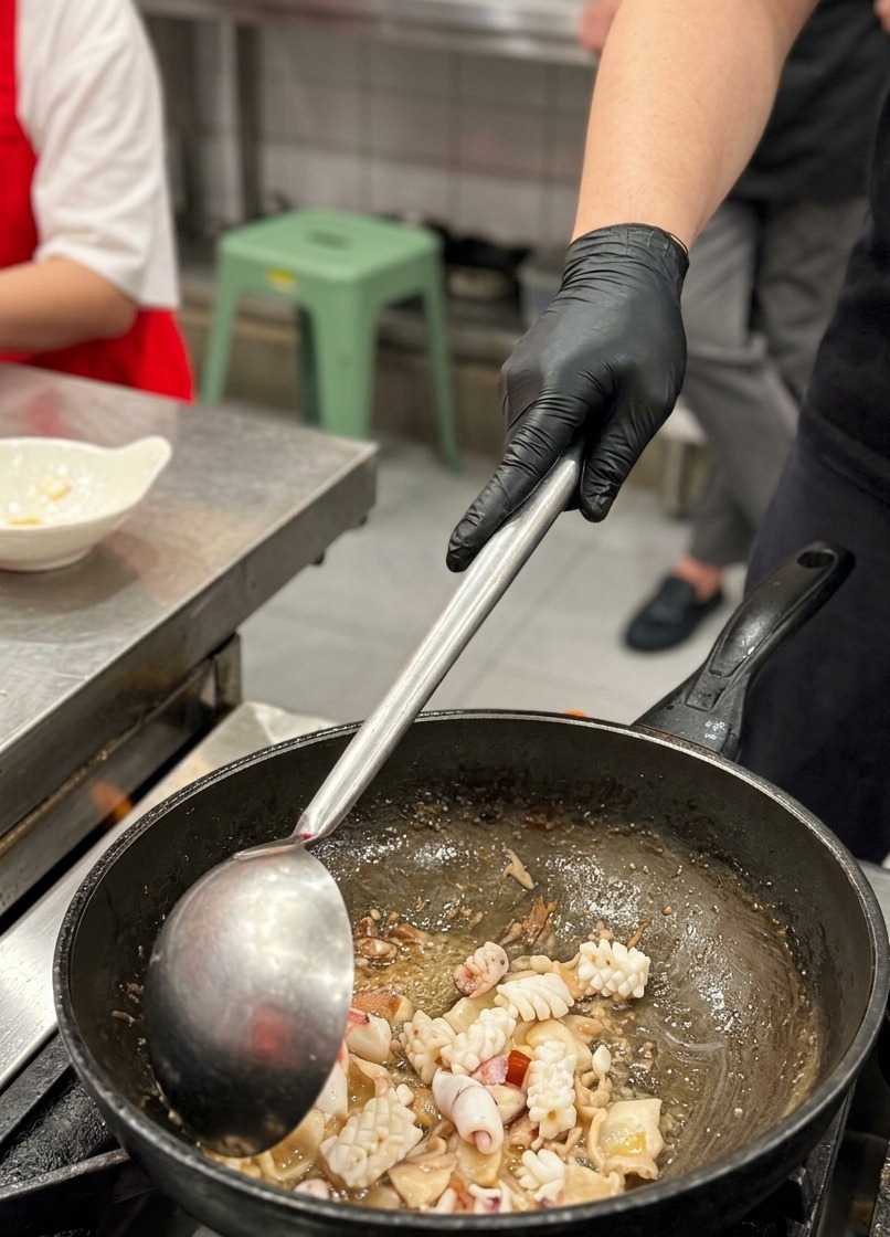 Primer plano de trozos de calamar con cortes en cuadrícula siendo integrados en un wok junto a tiras de panceta doradas usando un cucharón metálico.