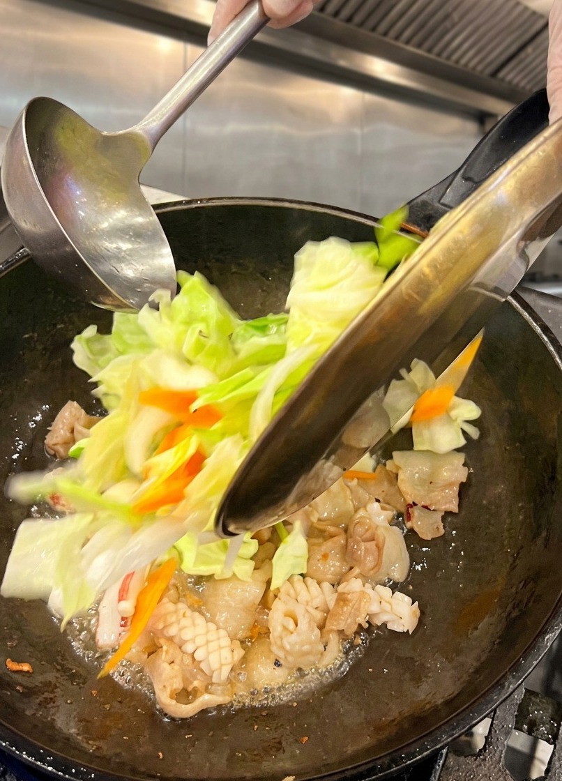 Repollo verde recién picado y rodajas de zanahoria siendo vertidos desde un plato de metal en un wok caliente que contiene panceta y calamar cocidos.