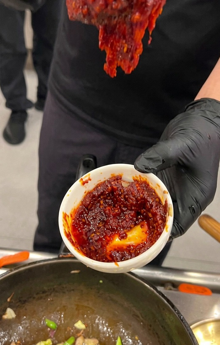 Una mano con guante negro sosteniendo un pequeño cuenco blanco lleno de una salsa de pasta de chile coreano roja y espesa sobre un wok de cocina.